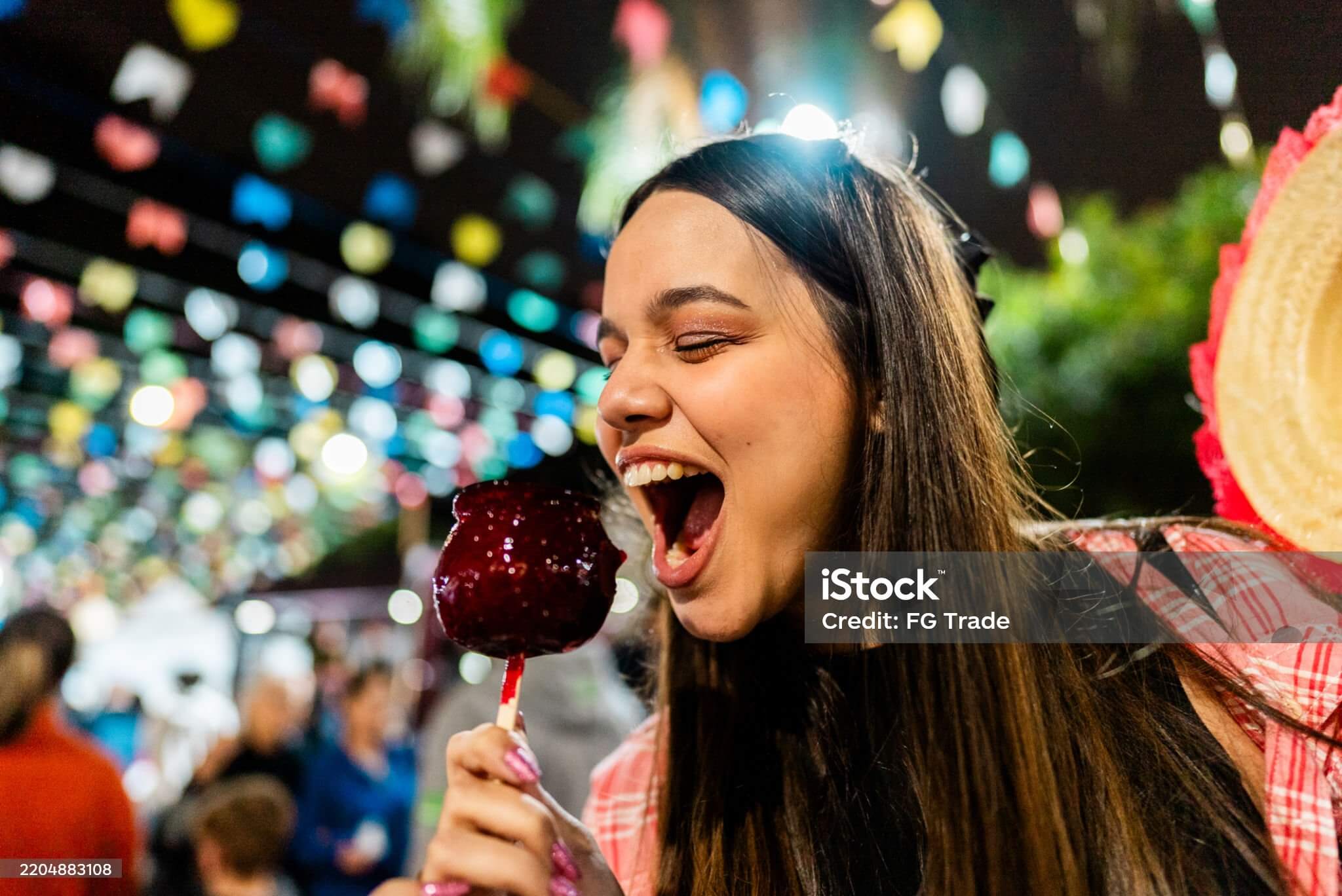 A girl with her mouth wide open ready to bite into a red toffee apple with a carnival scene in the background